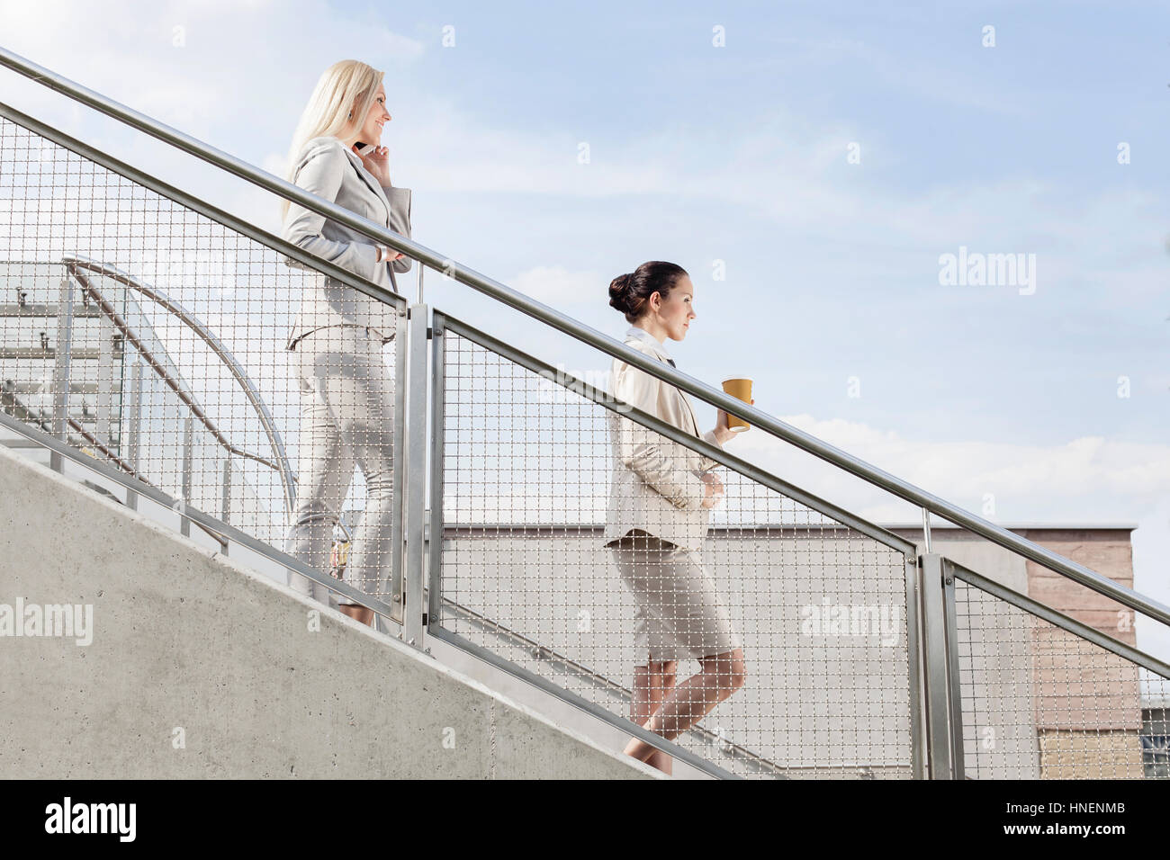 Profilo colpo di imprenditrici holding tazza monouso e il telefono cellulare mentre si sposta giù per le scale insieme contro sky Foto Stock