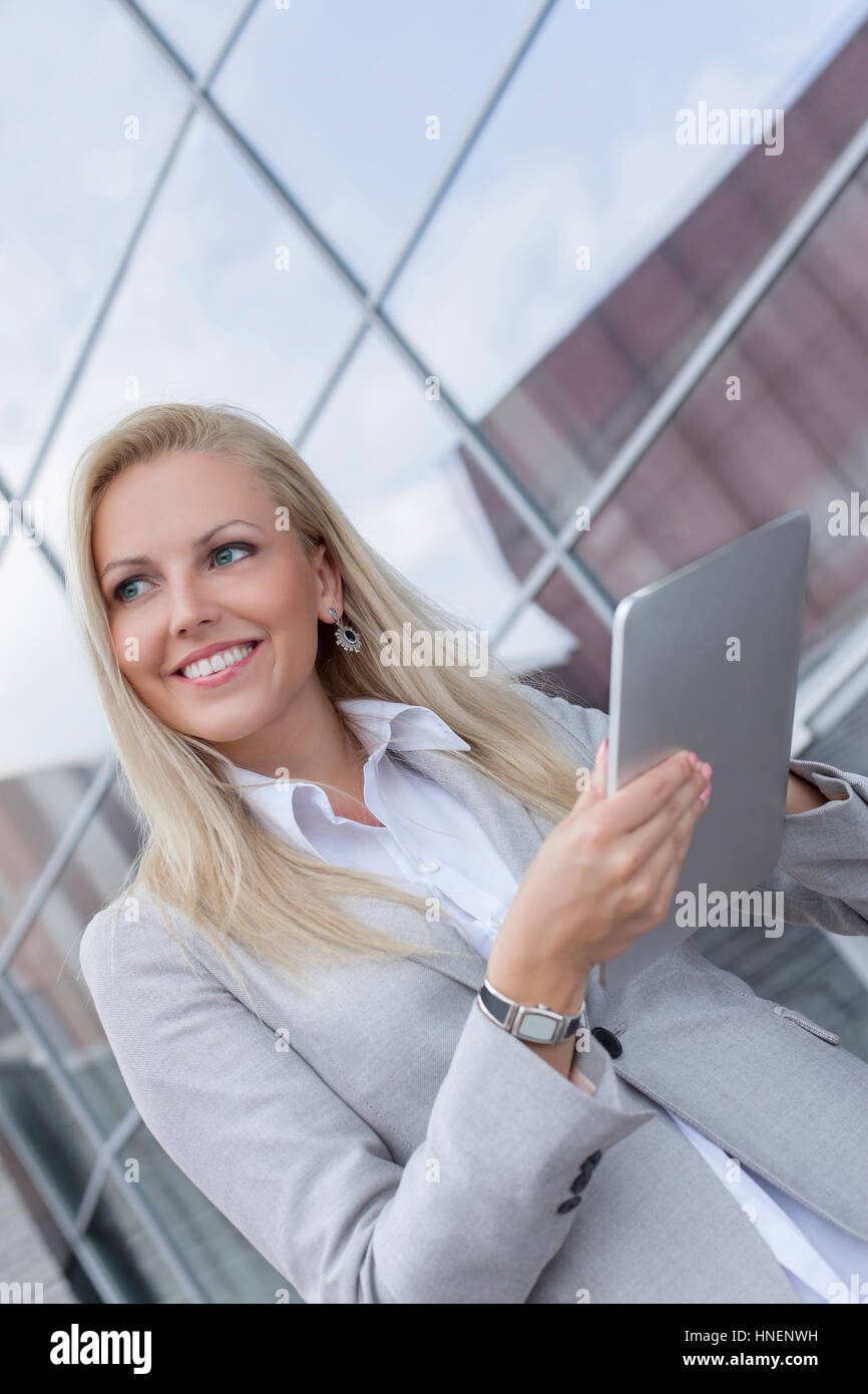 Felice imprenditrice guardando lontano tenendo digitale compressa contro l edificio per uffici Foto Stock