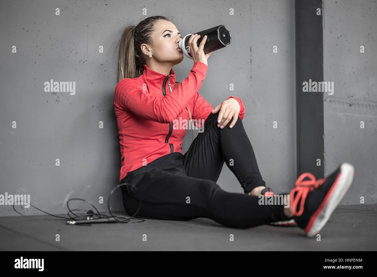 Attraente ragazza sportive si siede sul pavimento nella palestra accanto alla parete grigia. Ella beve dalla tazza nera nella mano destra e tiene la mano sinistra o Foto Stock