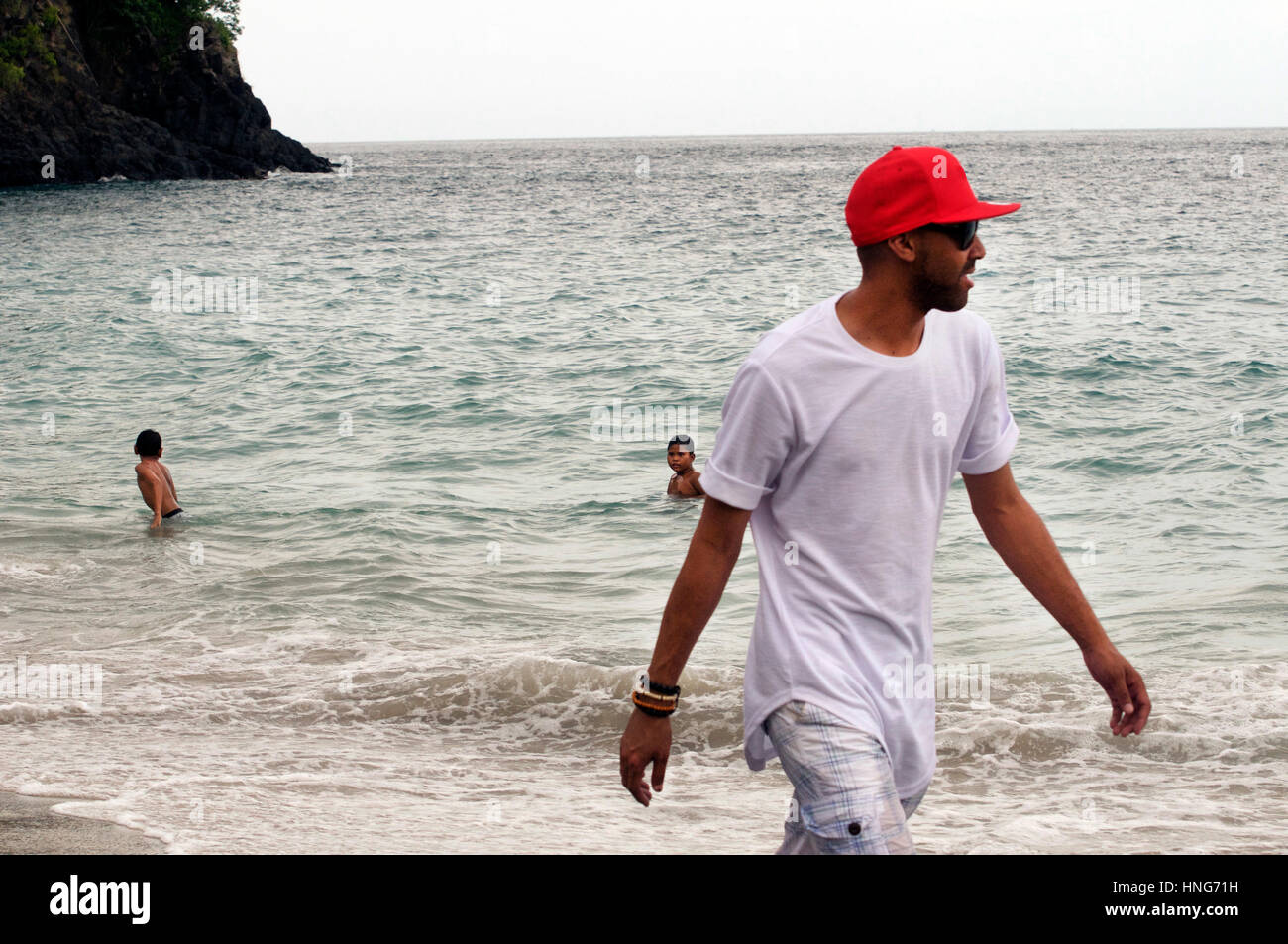 Cool Dude passeggiate lungo la spiaggia sabbiosa in Bali Indonesia indossando Red Baseball Hat, bianco maglietta e pantaloni corti Foto Stock