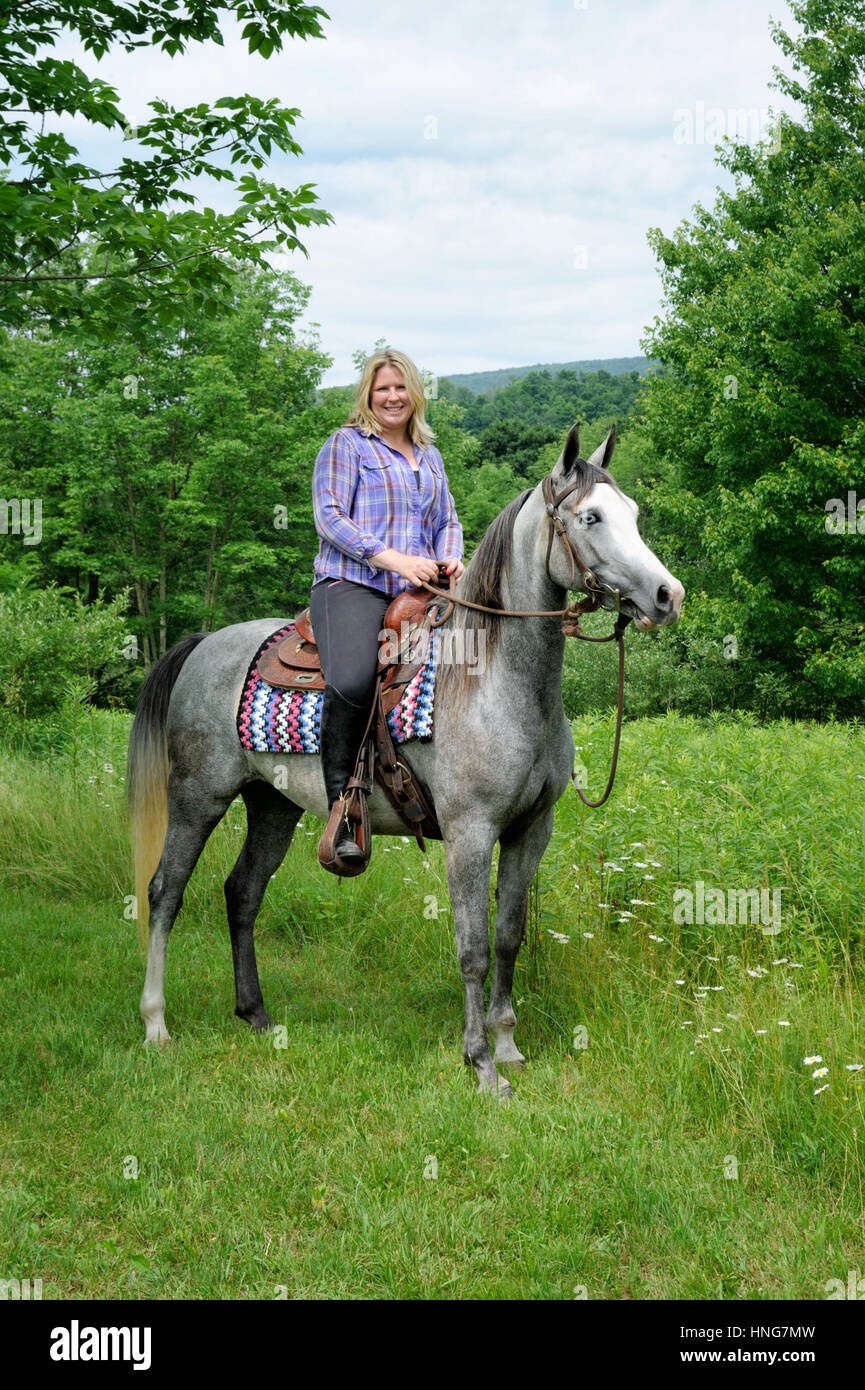 Bionda, donna in abiti casual, montato su un Blu-eyed, di fronte bianco-, pezzati cavallo, equitazione western in erba Campi verdi del fogliame di estate. PA Foto Stock