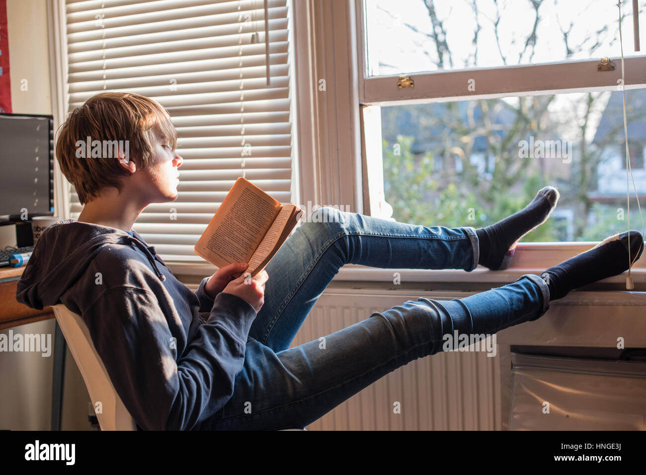 White ragazzo adolescente in jeans e un Sweat top udienza dal suo aprire la finestra della camera con i suoi piedi fino dalla sua scrivania di lettura e di revisione per gli esami GCSE Foto Stock