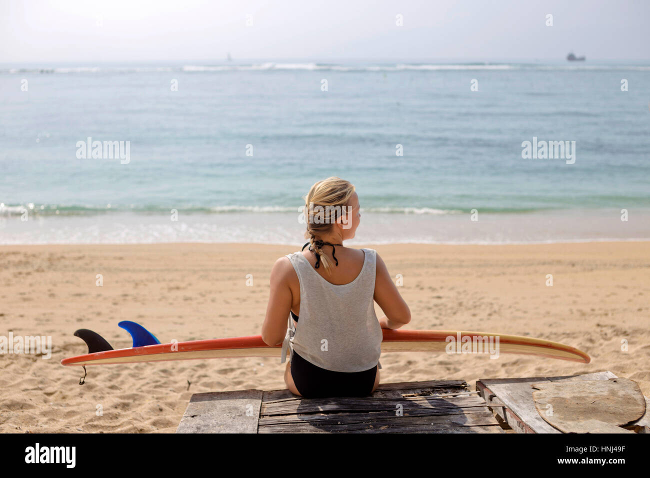 Vista posteriore della donna con la tavola da surf seduto alla spiaggia Foto Stock