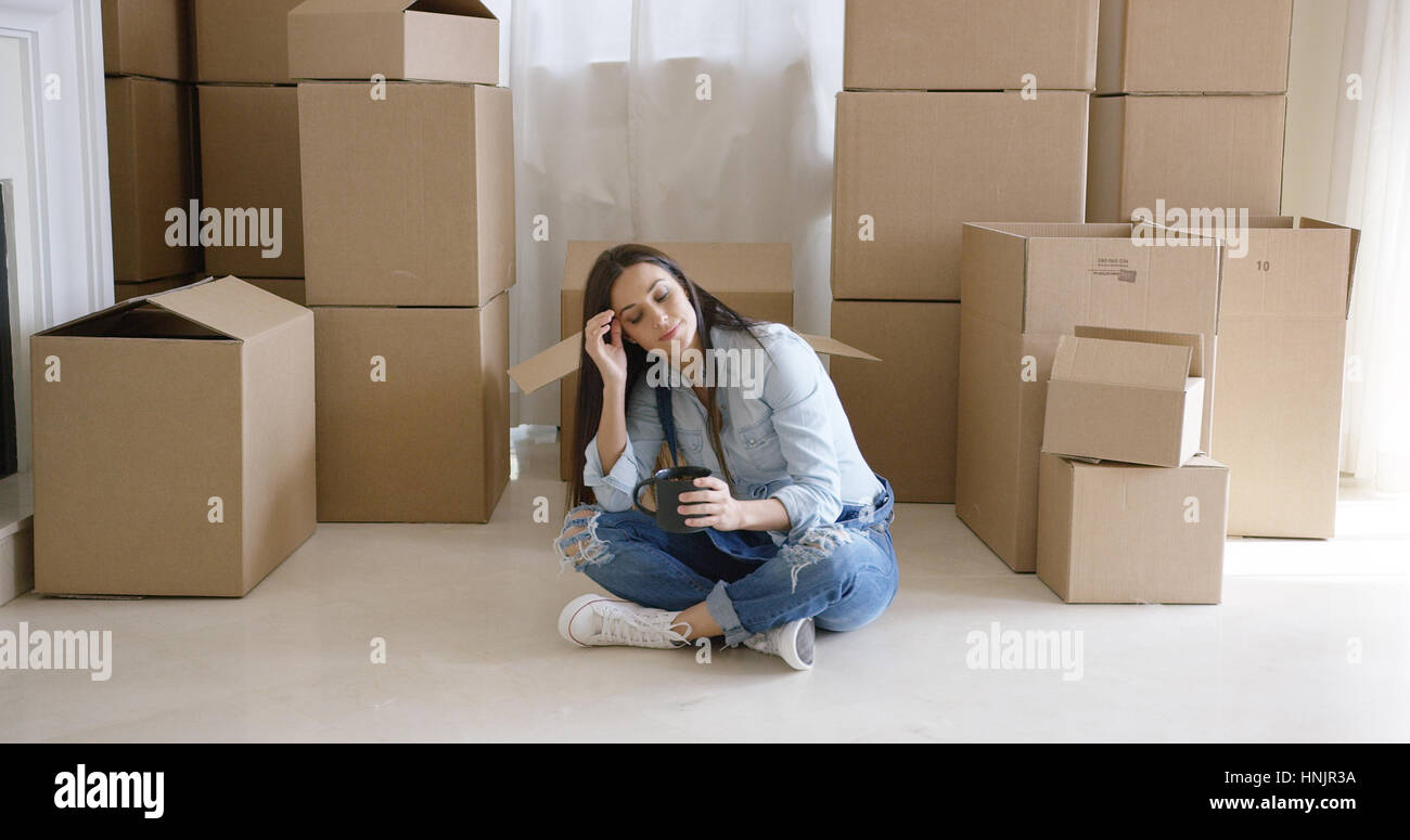 Stanco giovane donna prendendo una pausa dalla casa in movimento seduta con le gambe incrociate sul pavimento con caffè circondato dalle caselle Foto Stock