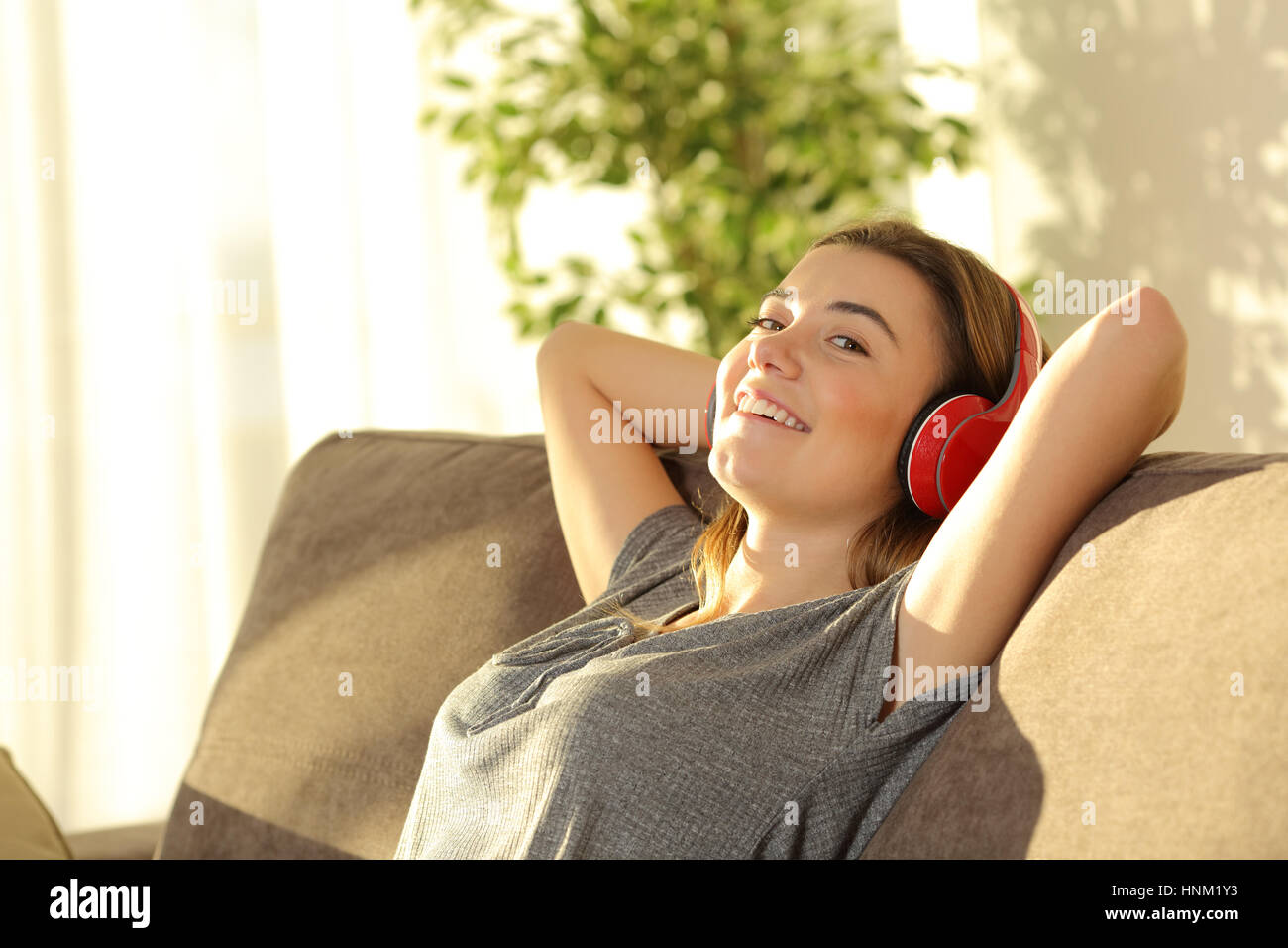 Un teenager di ascolto di musica con cuffie e guardando a voi seduto su un divano nel soggiorno di casa con una calda luce in background Foto Stock