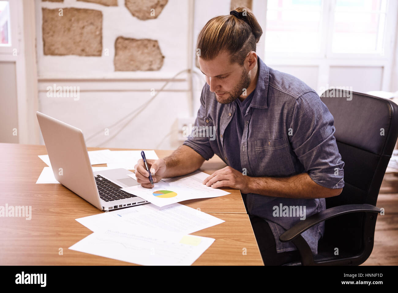 Giovane imprenditore con i suoi capelli legato indietro seduto alla sua scrivania con un computer portatile rendendo note su un grafico di fronte a lui con la mano destra Foto Stock