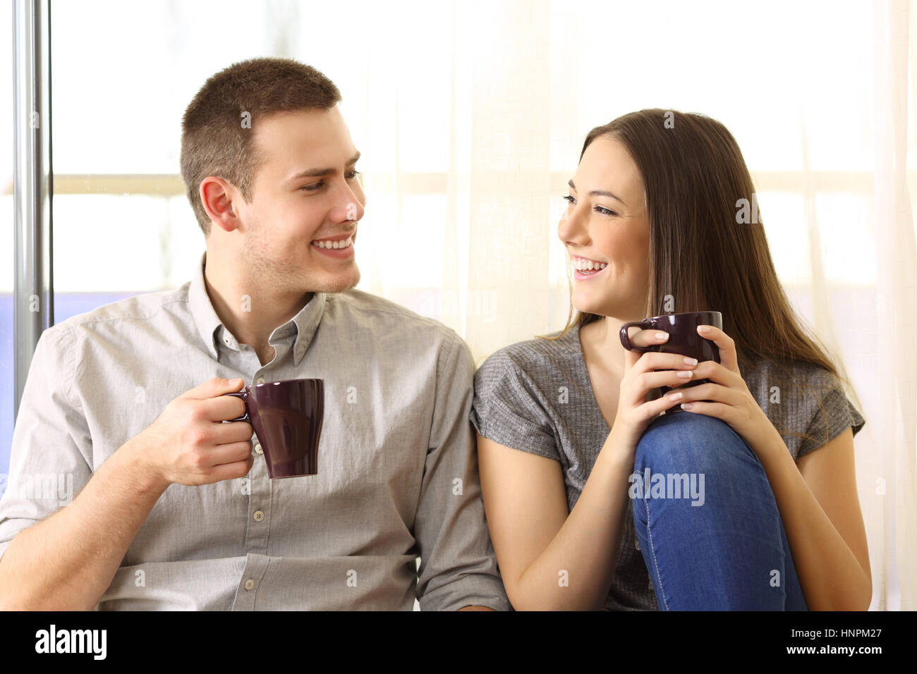 Vista frontale di una coppia felice di parlare e di bere il caffè seduta sul pavimento vicino a una finestra a casa Foto Stock