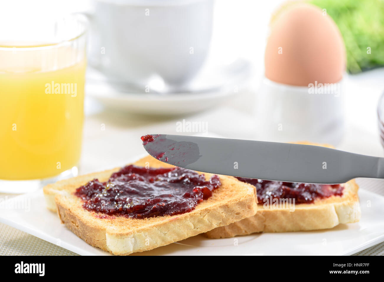 La deliziosa prima colazione con due toast su una piastra con marmellata, un coltello sulla parte anteriore e il succo di arancia con uovo dietro. Foto Stock
