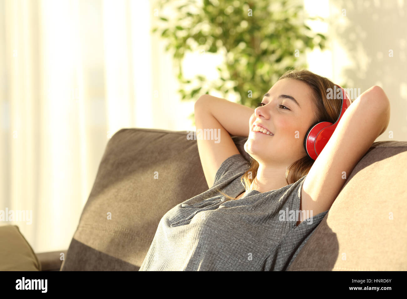 Un rilassato riposo teen e ascolto di musica con cuffie wireless seduto su un divano nel soggiorno di casa con una luce calda Foto Stock