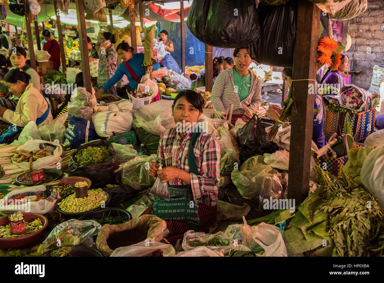 Femmina lavoratore di mercato nel mercato birmano circondato da prodotti alimentari guarda wistfully a telecamera con mercato generale attività in background Foto Stock