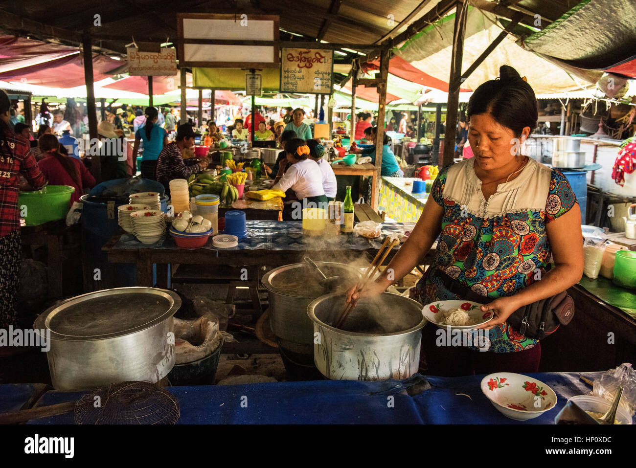 Donna birmano a Nyaung Shwe mercato, Myanmar che serve cibo dalla pentola grande in un ristorante con i clienti seduti in un ambiente colorato Foto Stock