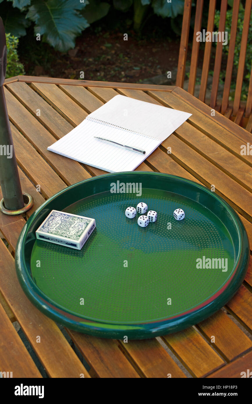 cinque dadi bianchi da 5 mm e mazzo di carte giacenti su un vassoio verde su un tavolo di legno con un notebook e una penna accanto, pronti per i giocatori per iniziare la partita Foto Stock