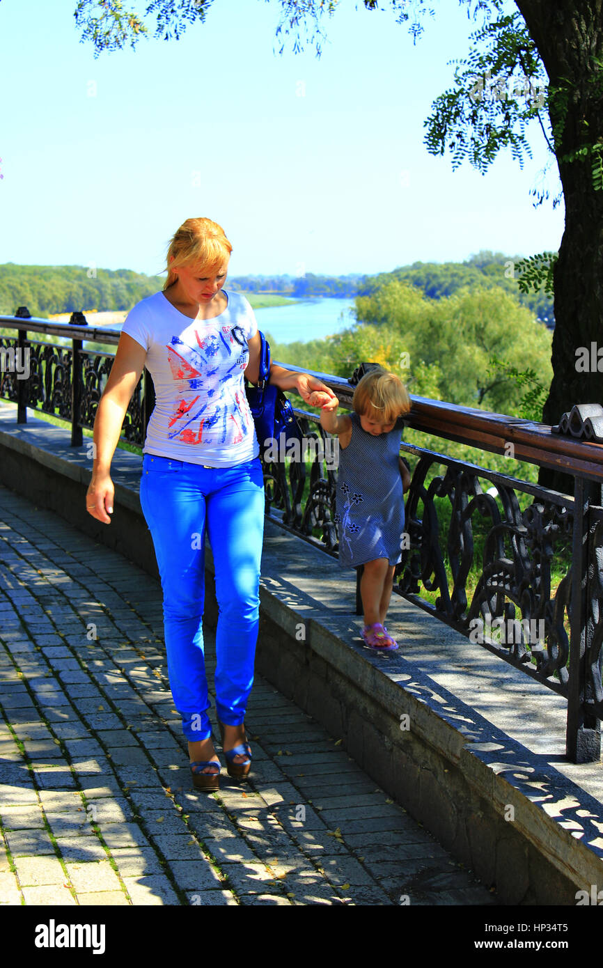 Moderna moda donna conduce la sua piccola figlia per mano sul cordolo in un parco della città Foto Stock