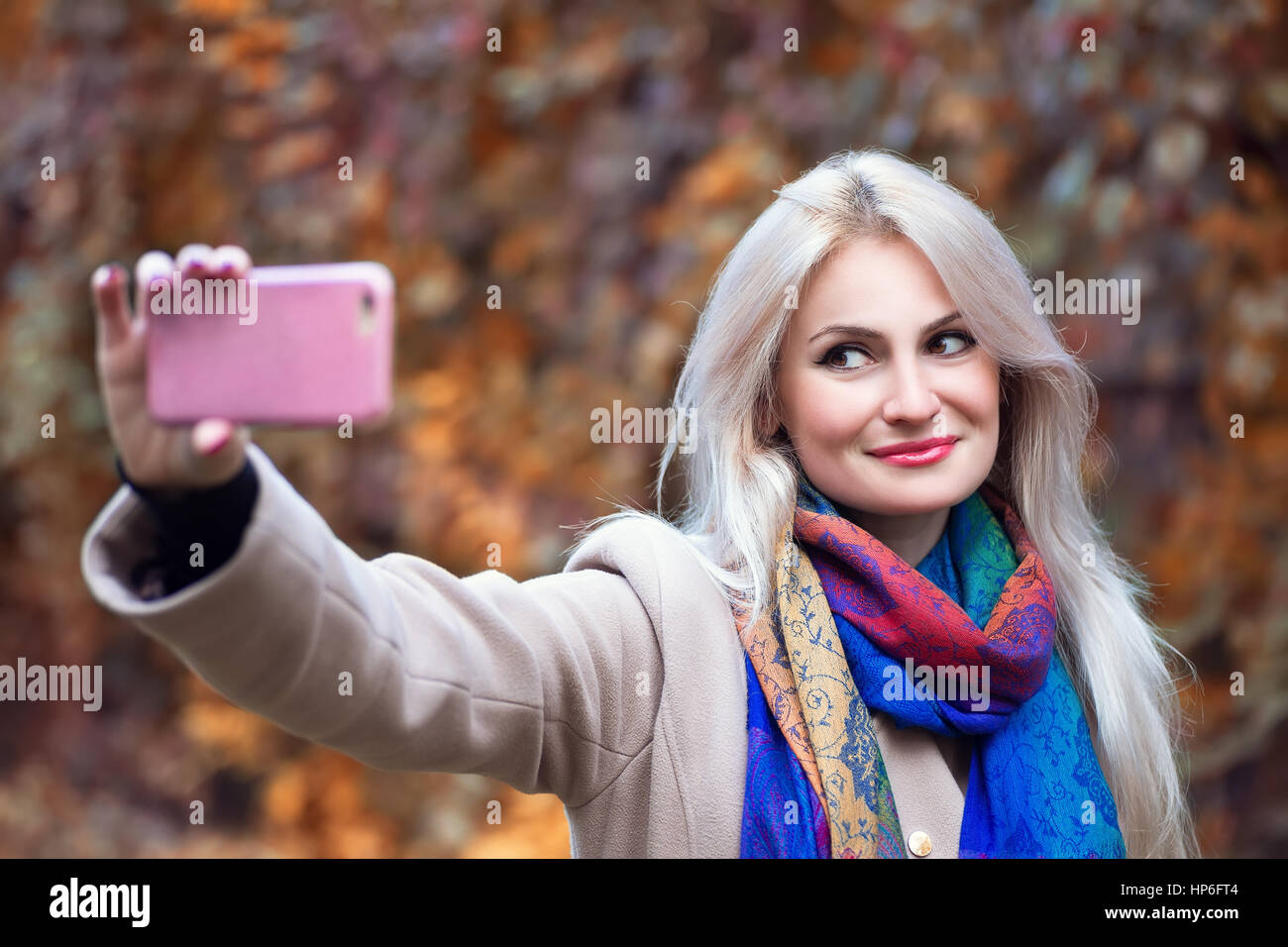 Bella bionda e giovane donna caucasica tenendo selfie con lo smartphone all'aperto nel parco in autunno. Selfie autunno. In autunno, persone, tecnologia, stile di vita Foto Stock