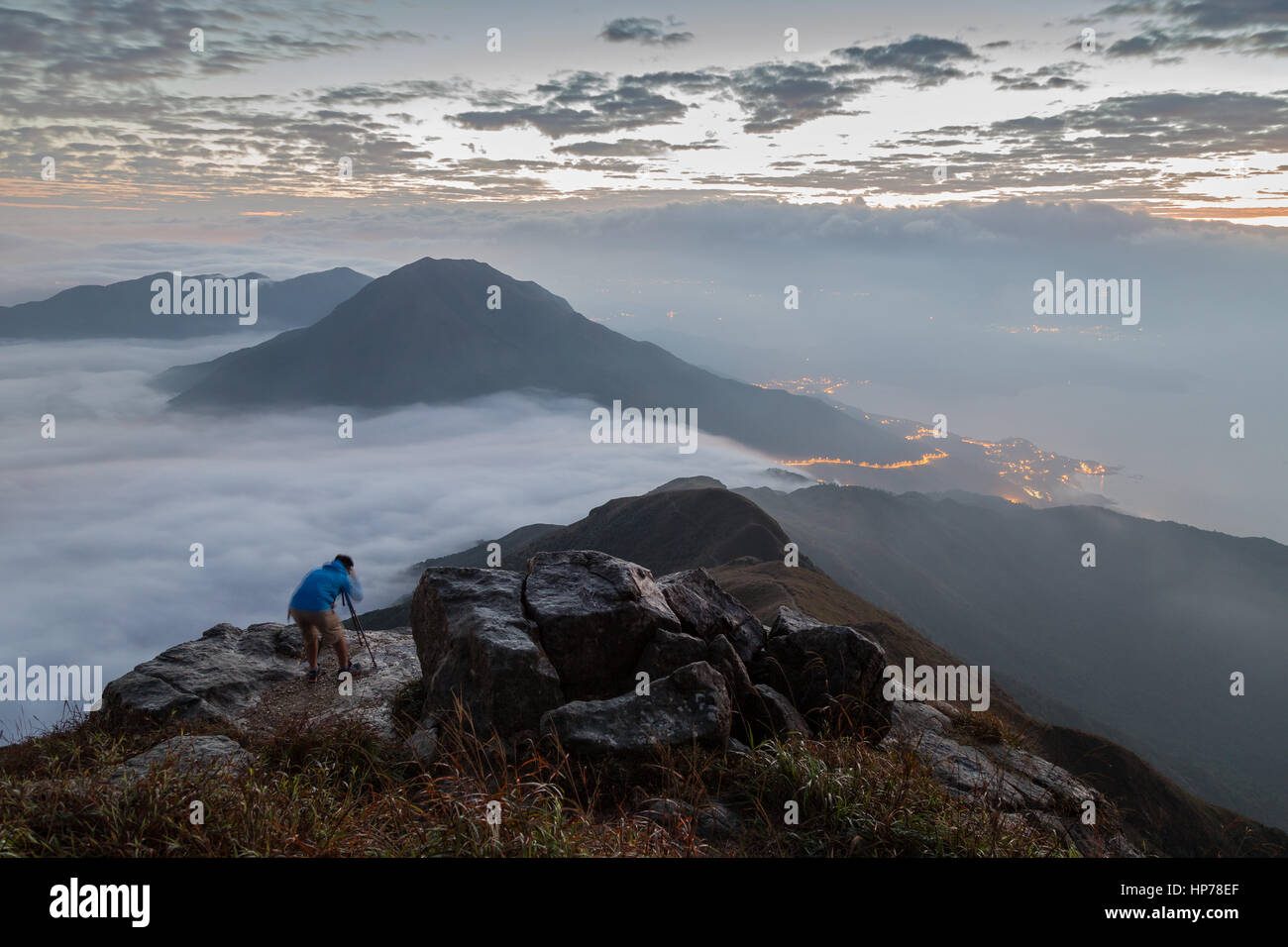 Vista di nuvole, cime, costa e un uomo con una videocamera e treppiede in piedi su una roccia a picco di Lantau sull'Isola di Lantau in Hong Kong all'alba Foto Stock