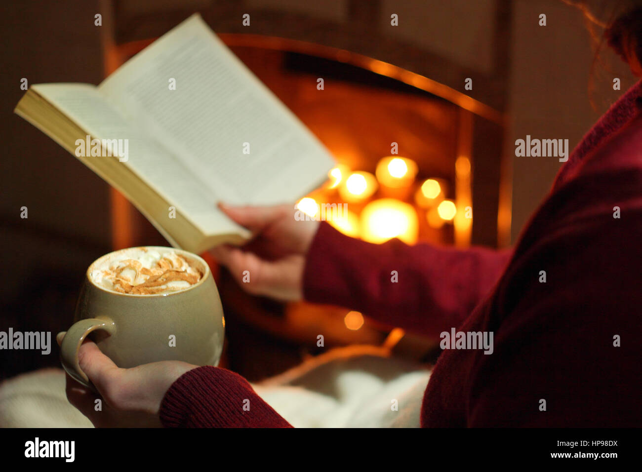 Una donna che legge un libro con una tazza di panna e rabboccato mocha (caffè e cioccolato) da un caminetto acceso con candele in un accogliente English home in inverno Foto Stock