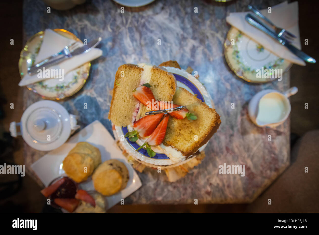Tè pomeridiano. Un overhead immagine della torta decorata con fragole, focaccine, latte, piatti e posate su un tavolo di marmo. Foto Stock