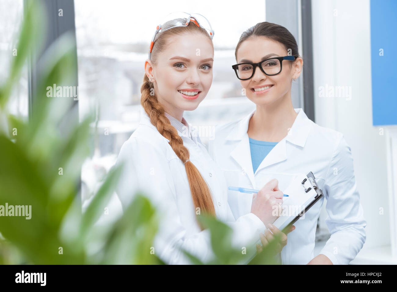 Le giovani ricercatrici in camice da laboratorio e bicchieri prendendo appunti e sorridente in telecamera Foto Stock