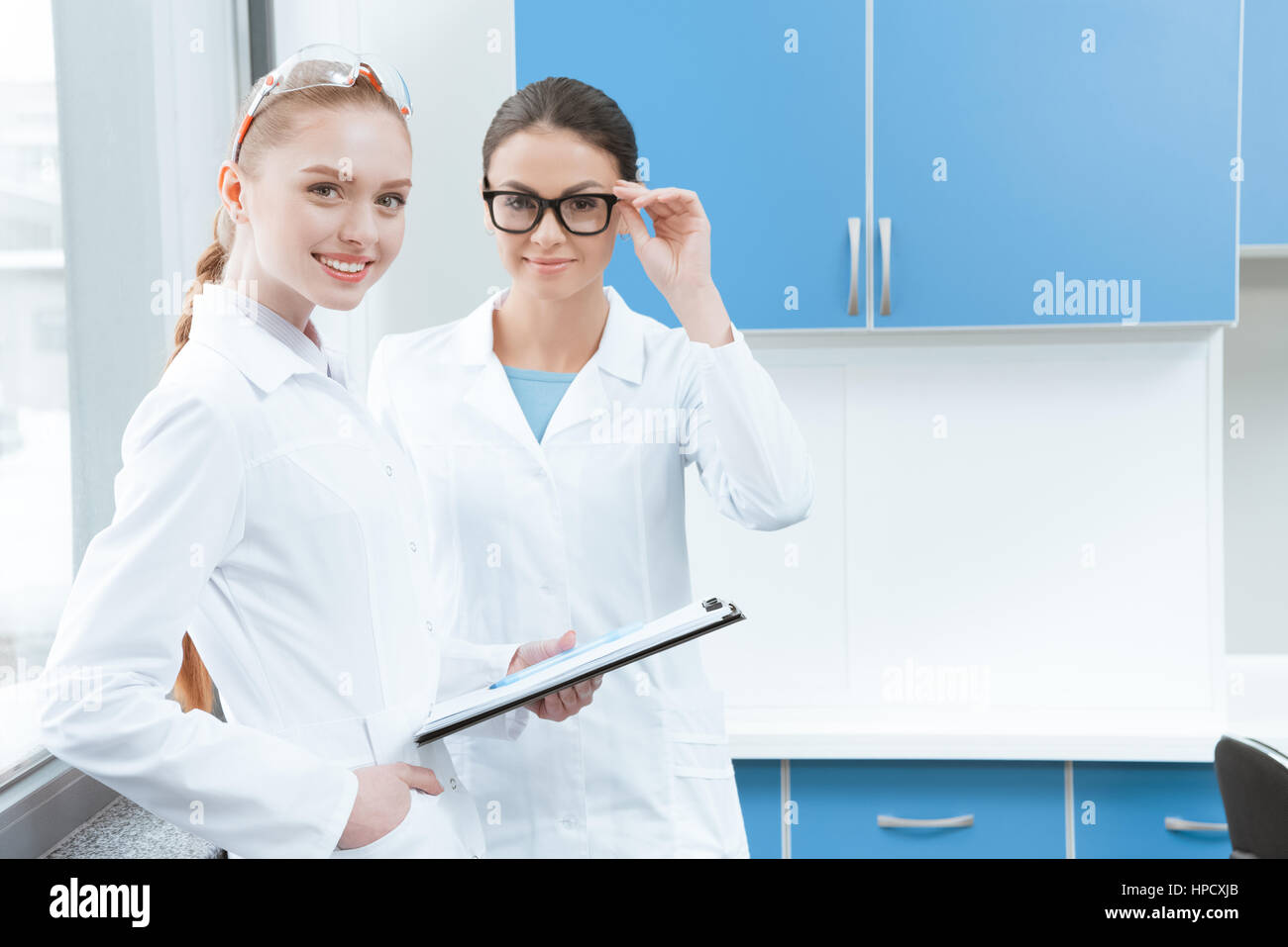 Le giovani ricercatrici in camici ed occhiali sorridente in telecamera in laboratorio Foto Stock