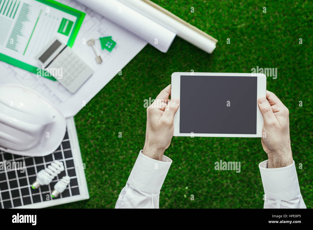 Progetto di casa, pannello solare, casco su erba lussureggiante e maschio di mani tenendo un tablet, verde costruzione sostenibile e le energie alternative concept Foto Stock