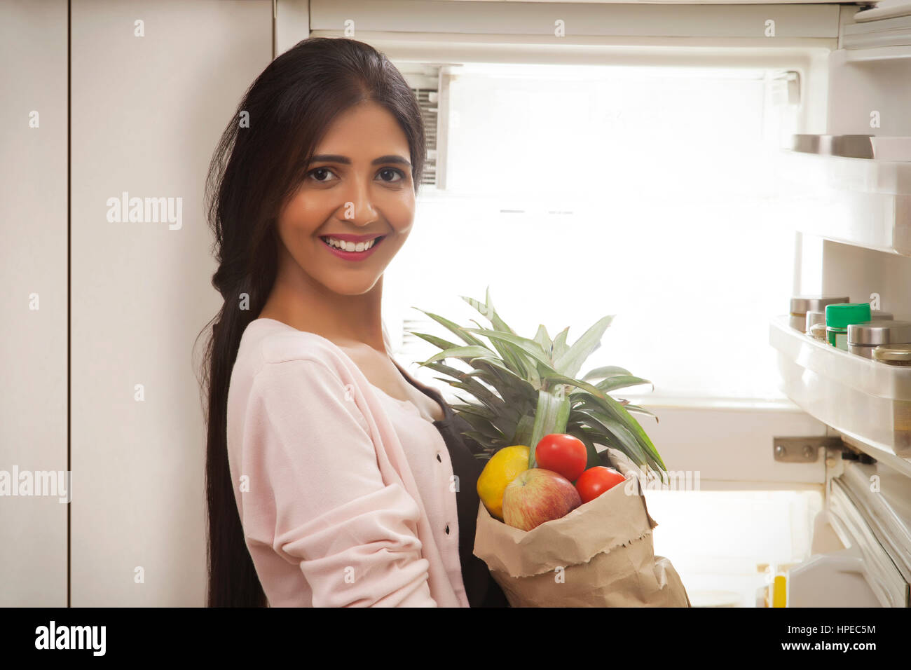 Sorridente giovane donna azienda sacchetto di frutti in piedi nella parte anteriore del frigorifero aperto Foto Stock