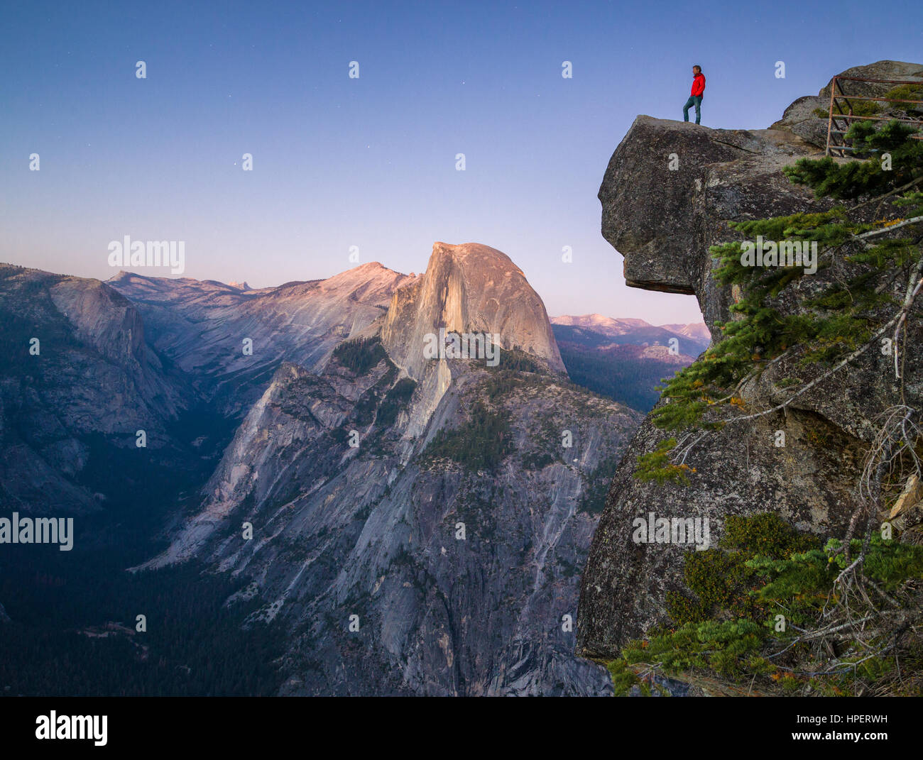 Un intrepido escursionista è in piedi su una roccia a strapiombo godendo della vista verso il famoso Half Dome presso il Glacier Point si affacciano in Twilight, Yosemite NP, STATI UNITI D'AMERICA Foto Stock