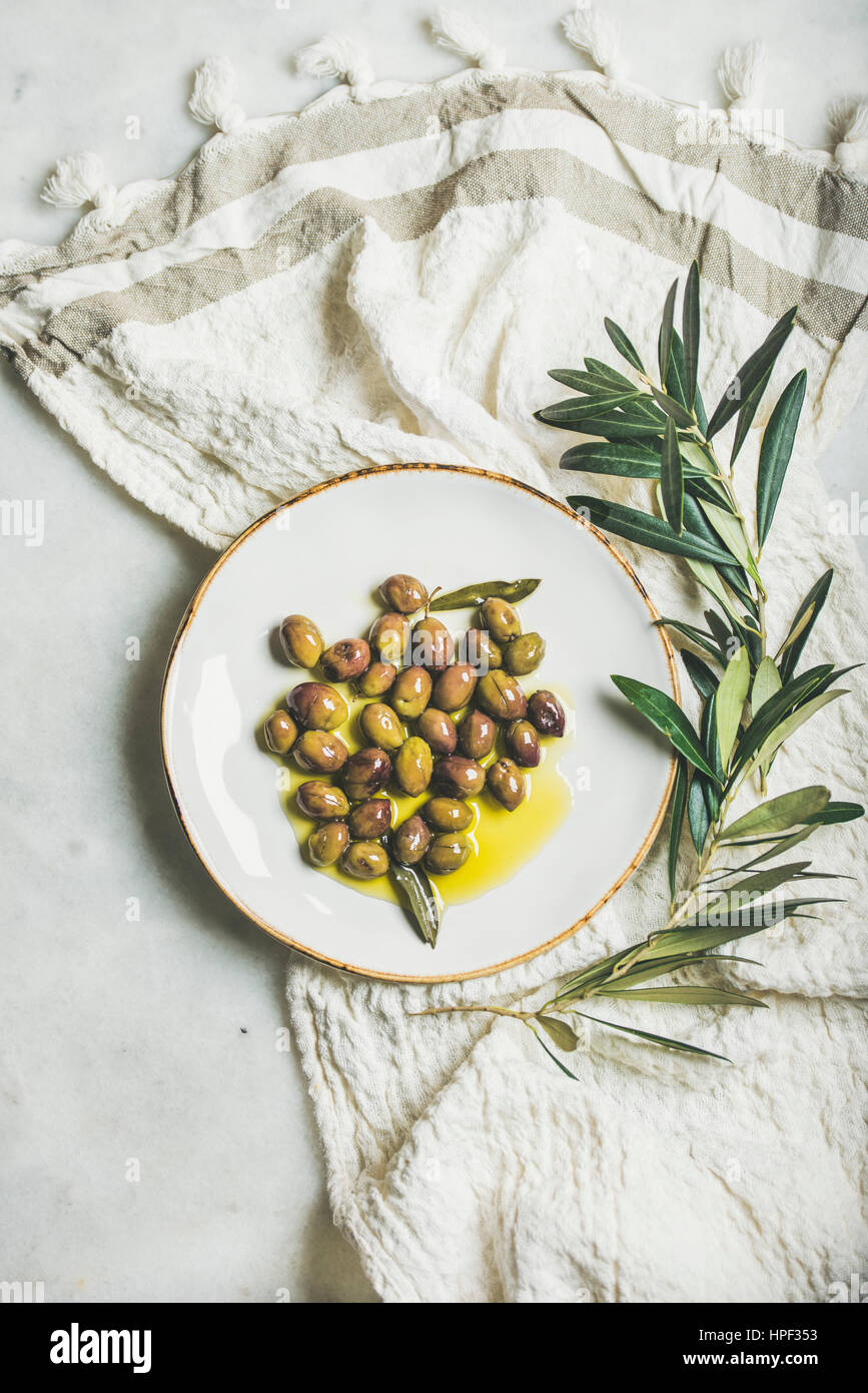 Decapare il verde mediterraneo delle olive in olio vergine di oliva su bianco piastra ceramica e ulivo ramo marmo grigio Sfondo, vista dall'alto Foto Stock