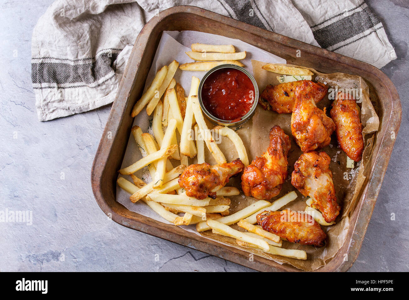 Il fast food fritto di pollo piccante gambe, ali e le patatine fritte di patate con sale e ketchup servita sulla carta da forno nel vecchio forno il vassoio con carta asciugatutto o Foto Stock