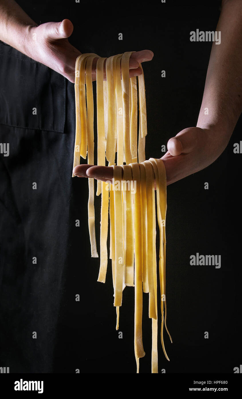 Crudo fresco non cotti pasta fatta in casa tagliatelle in mani dell'uomo oltre il grembiule nero come sfondo. Foto Stock