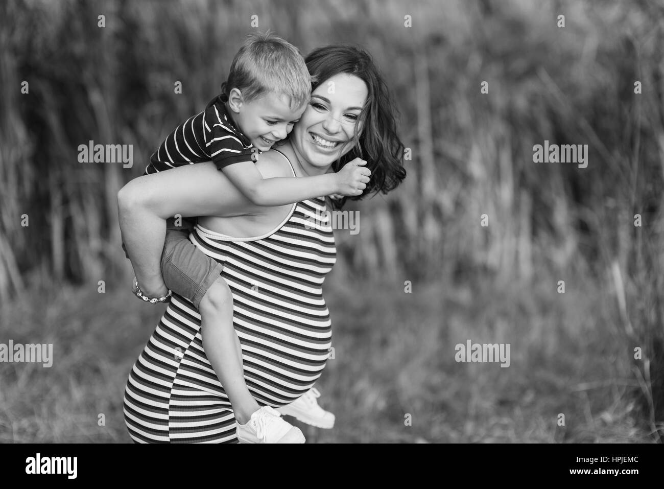 Bambino seduto sulle spalle di sua madre in stato di gravidanza. Felice madre e suo figlio. Sfondo verde sfumato, all'esterno. Foto in bianco e nero. Foto Stock