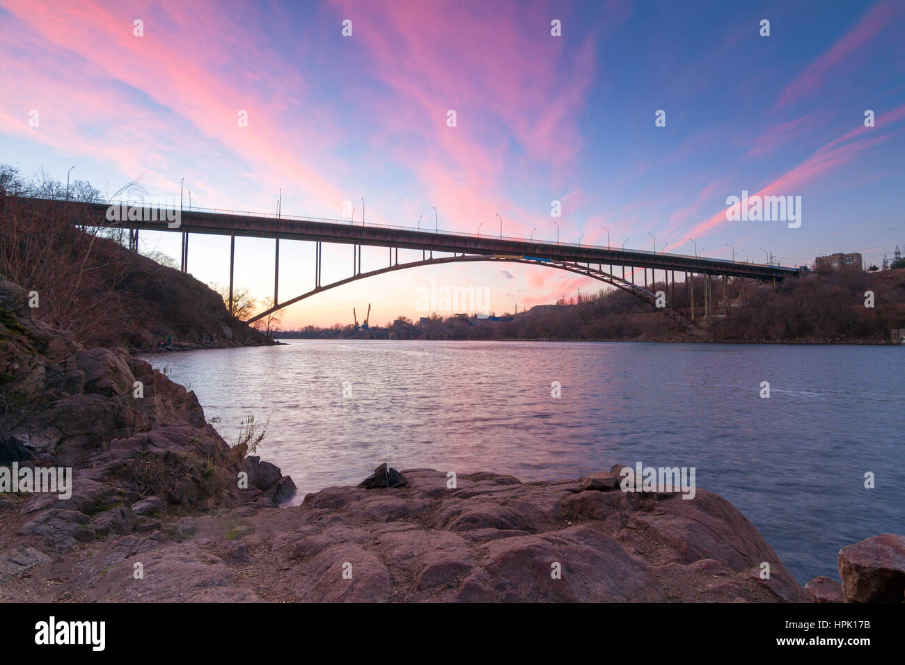 Acciaio ponte ad arco sul vecchio Dnieper con luminosi nuvole nel cielo della sera riflettendo in acqua e antiche rocce di granito Khortytsia island, Zaporozhye, Ukr Foto Stock