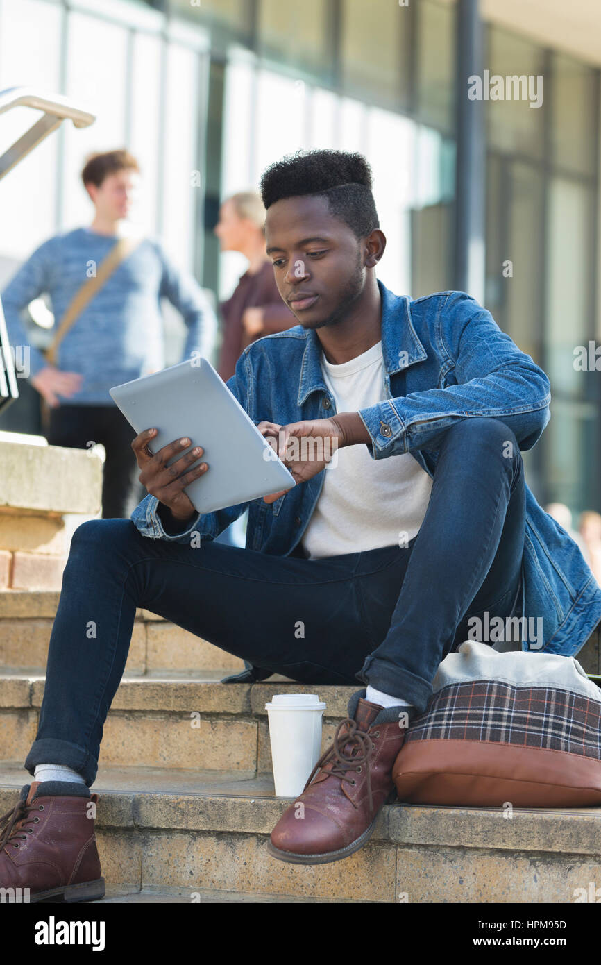 Un felice giovane studente maschio sorrisi come egli studi all'esterno. Egli sta cercando la sua tavoletta digitale e ha una bevanda calda e la sua borsa da i suoi piedi. Foto Stock