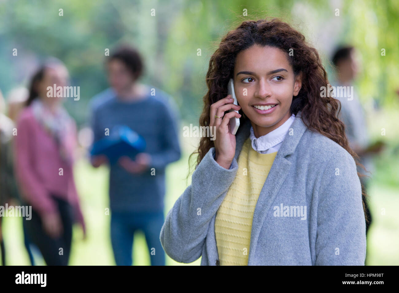 Un felice giovane studente sorrisi come usa il suo telefono cellulare all'esterno. Essa è avvolta in un rivestimento di spessore. Un gruppo di studenti che sono al di fuori della messa a fuoco in bac Foto Stock