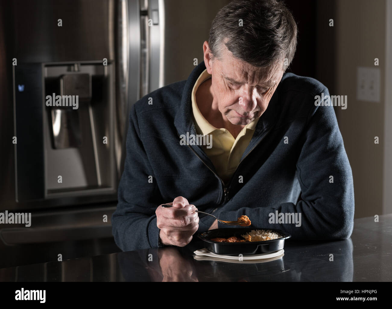 Solitario e depresso maschio senior seduto da solo al tavolo della cucina di mangiare un cotto in forno a microonde piatto pronto di curry dal vassoio di plastica Foto Stock