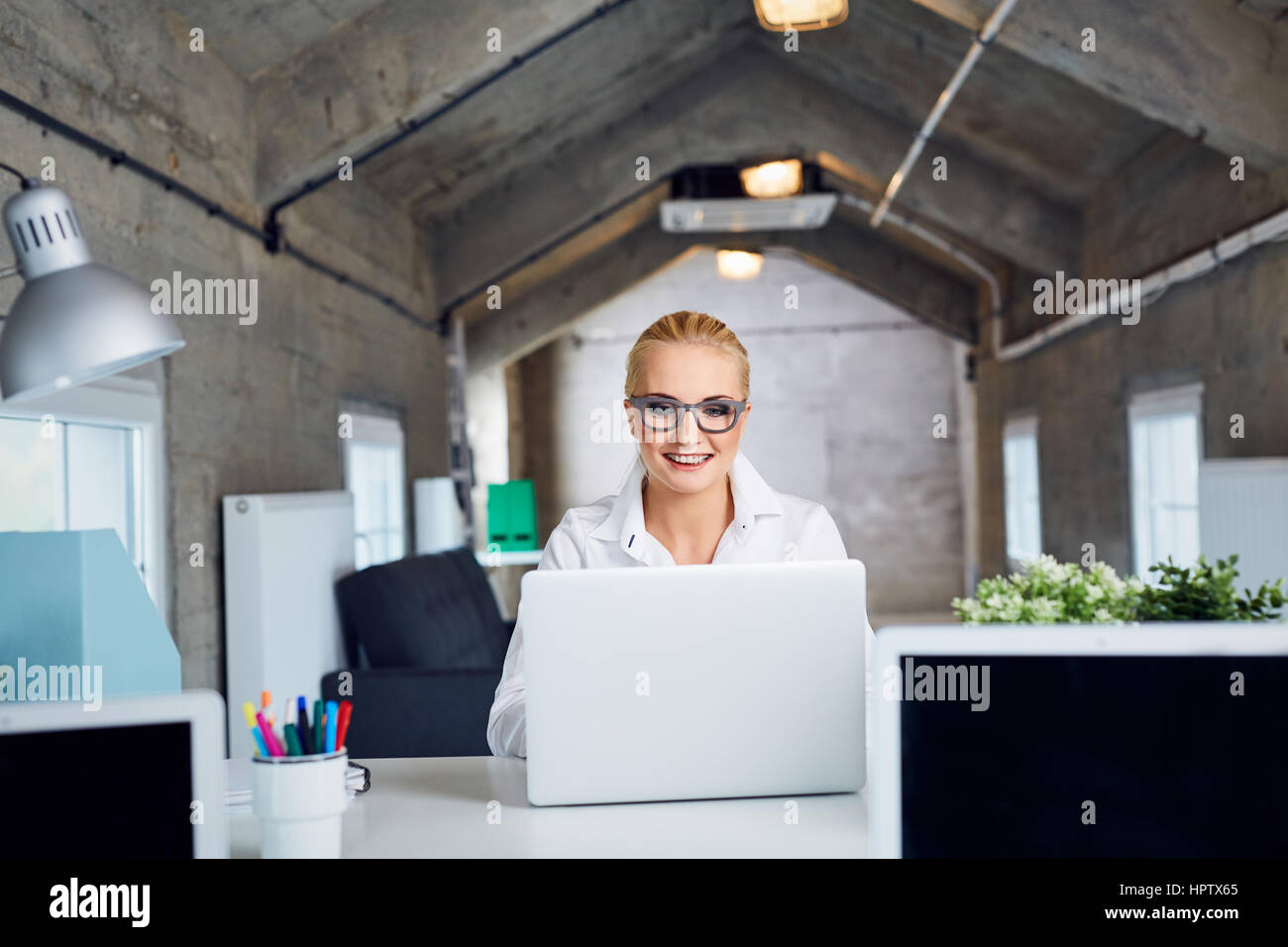 Allegro giovane donna con gli occhiali a sorridere mentre si lavora sul computer portatile in ufficio moderno Foto Stock