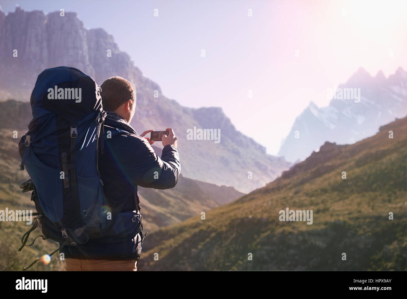 Giovane uomo con zaino tramite telefono cellulare dotato di fotocamera digitale nella soleggiata valle sotto le montagne Foto Stock