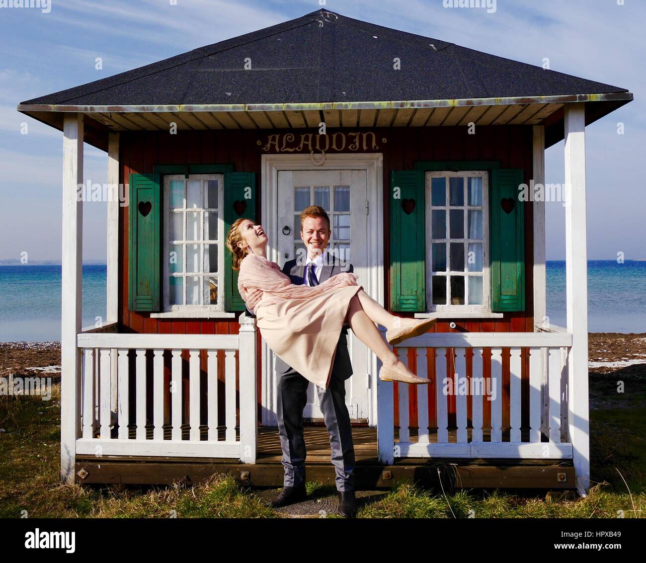 Un attraente felice giovane marito portando la sua bella moglie di nuovo al di sopra della soglia del simpatico, colorato Beach Hut per divertimento sull'isola danese di Aero Foto Stock