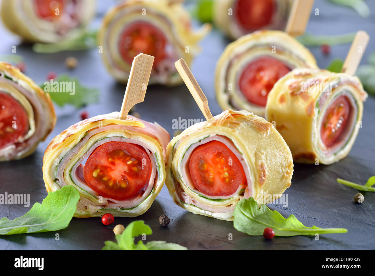 Deliziosi stuzzichini: rotoli di frittelle sottili (crepes) con prosciutto, formaggio spalmabile, insalata di rucola e pomodori ciliegia servita su un ardesia Foto Stock