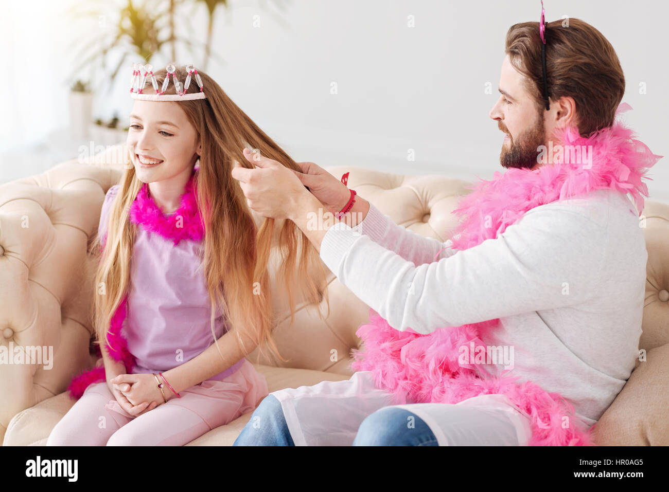 Tempo di bellezza. Bel uomo barbuto mantenendo piuma rosa intorno al suo collo indossando ponticello bianco tenendo i capelli di sua figlia nelle sue mani Foto Stock