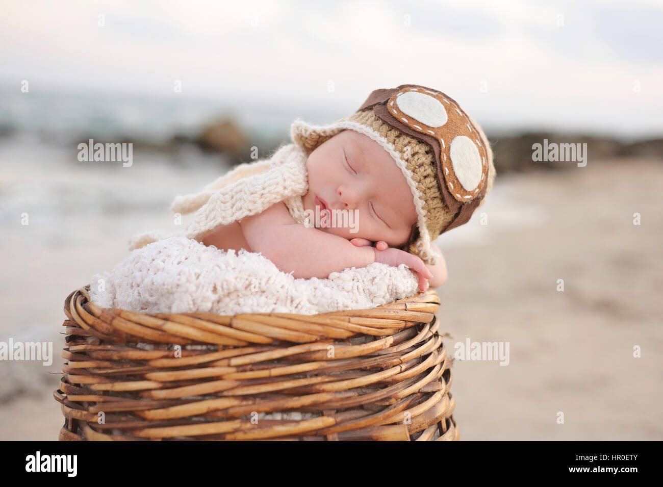 Due settimane vecchio neonato Bambino addormentato in un cesto di vimini indossando un aviatore hat. Fotografato su una spiaggia con una roccia jetty. Foto Stock