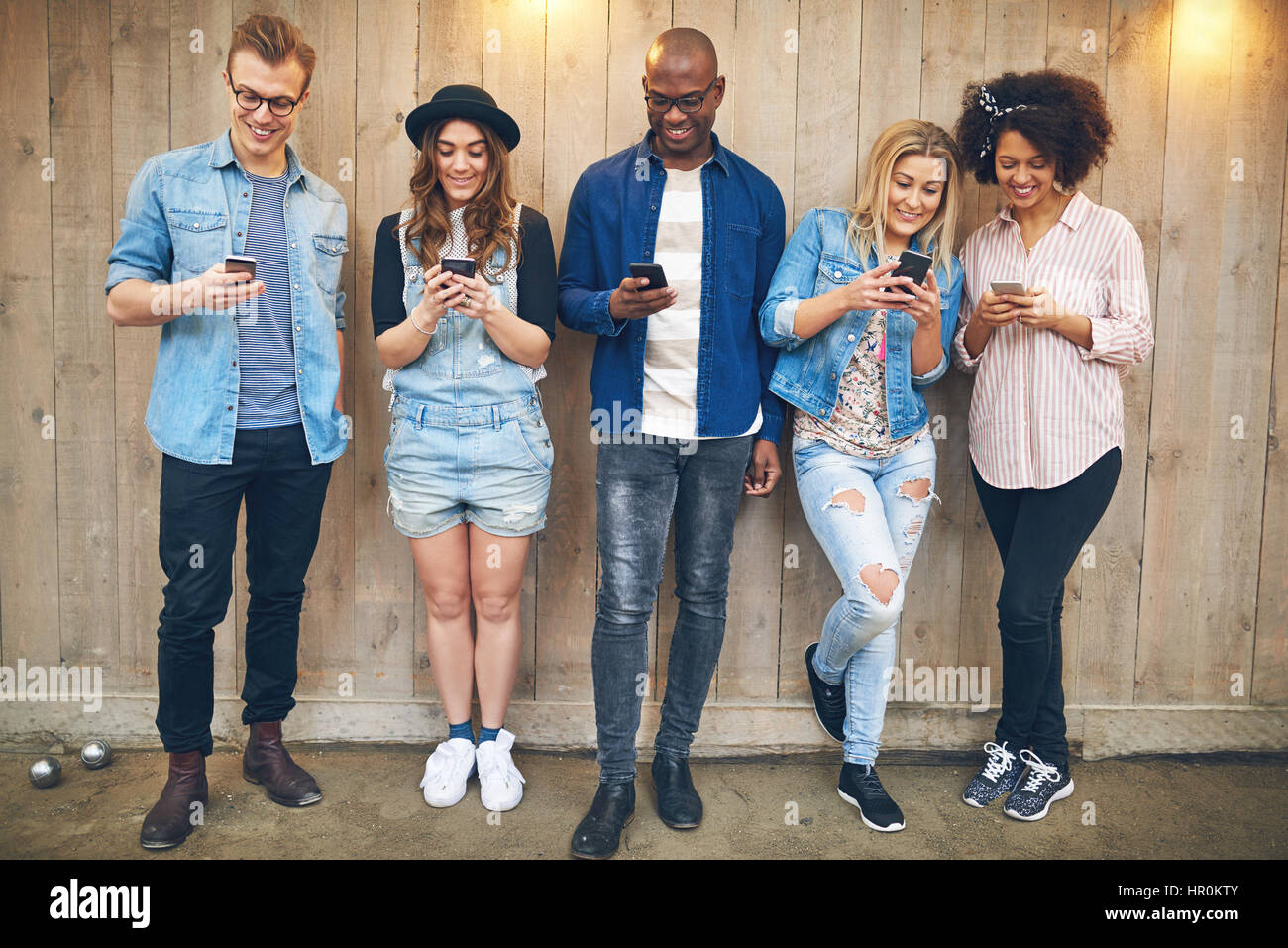 Un gruppo di giovani a parte o sale riunioni di indossare abiti casual in piedi vicino alla parete di legno tutti staring al loro smartphone, tutti sorridenti Foto Stock