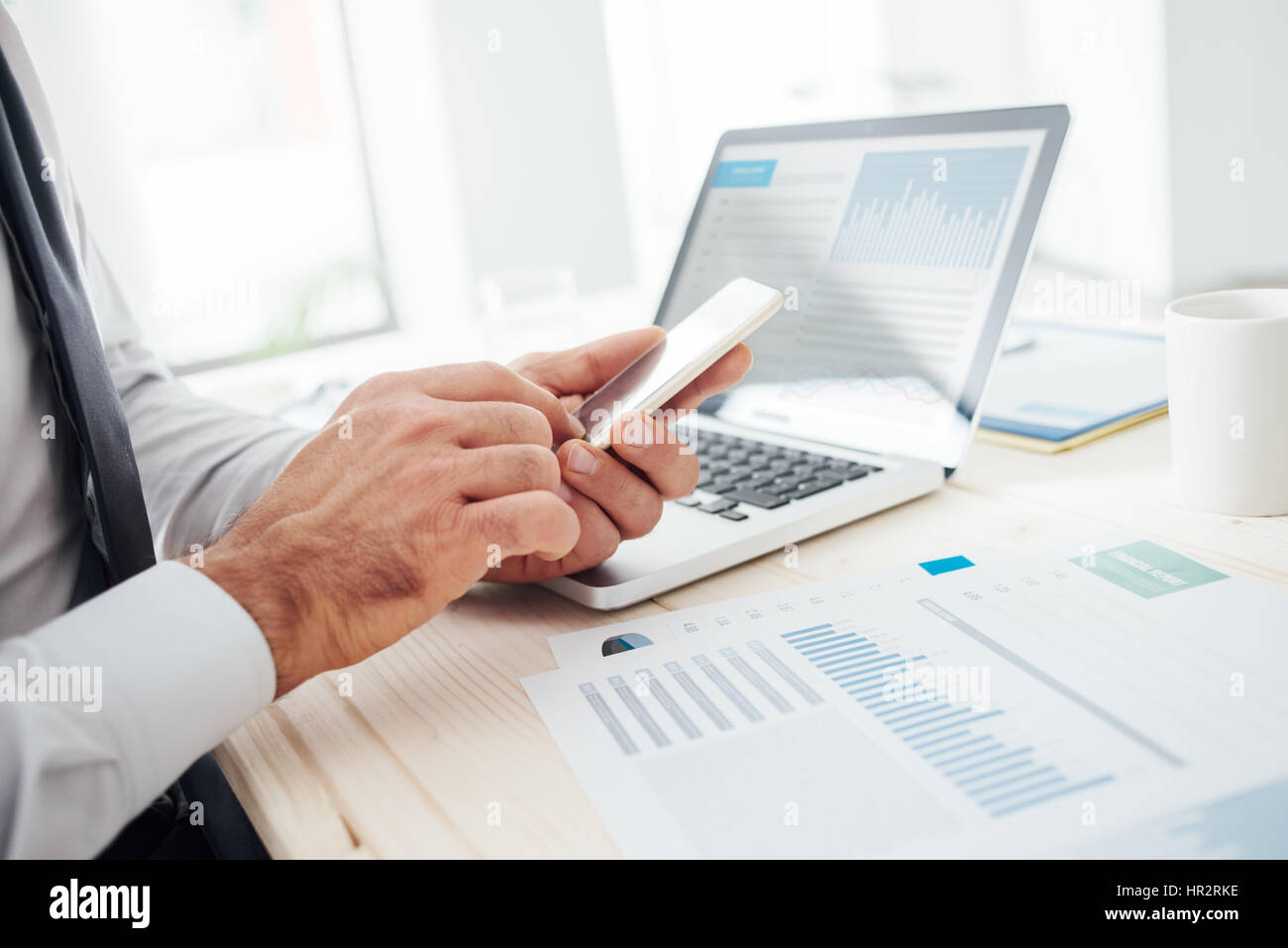 Imprenditore al desk utilizzando un'applicazione finanziaria sul suo smart phone e lavorare sulle relazioni e le mani vicino fino Foto Stock