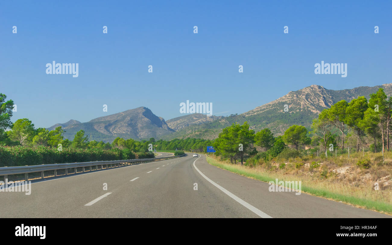 Sole sulla autostrada costiera. Lone bianco auto trascina lungo attraverso colline e le gamme della montagna sui bordi dell'Europa continentale in Spagna rurale. Foto Stock