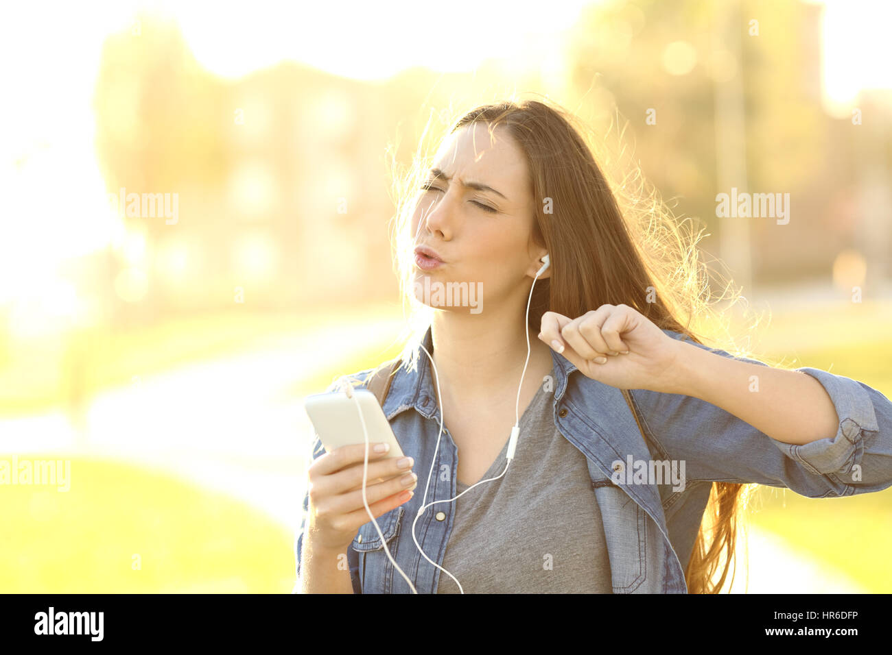 Donna allegra ascoltando canzoni da uno smart phone e sentire la musica in strada al tramonto con una luce calda del sole in background Foto Stock