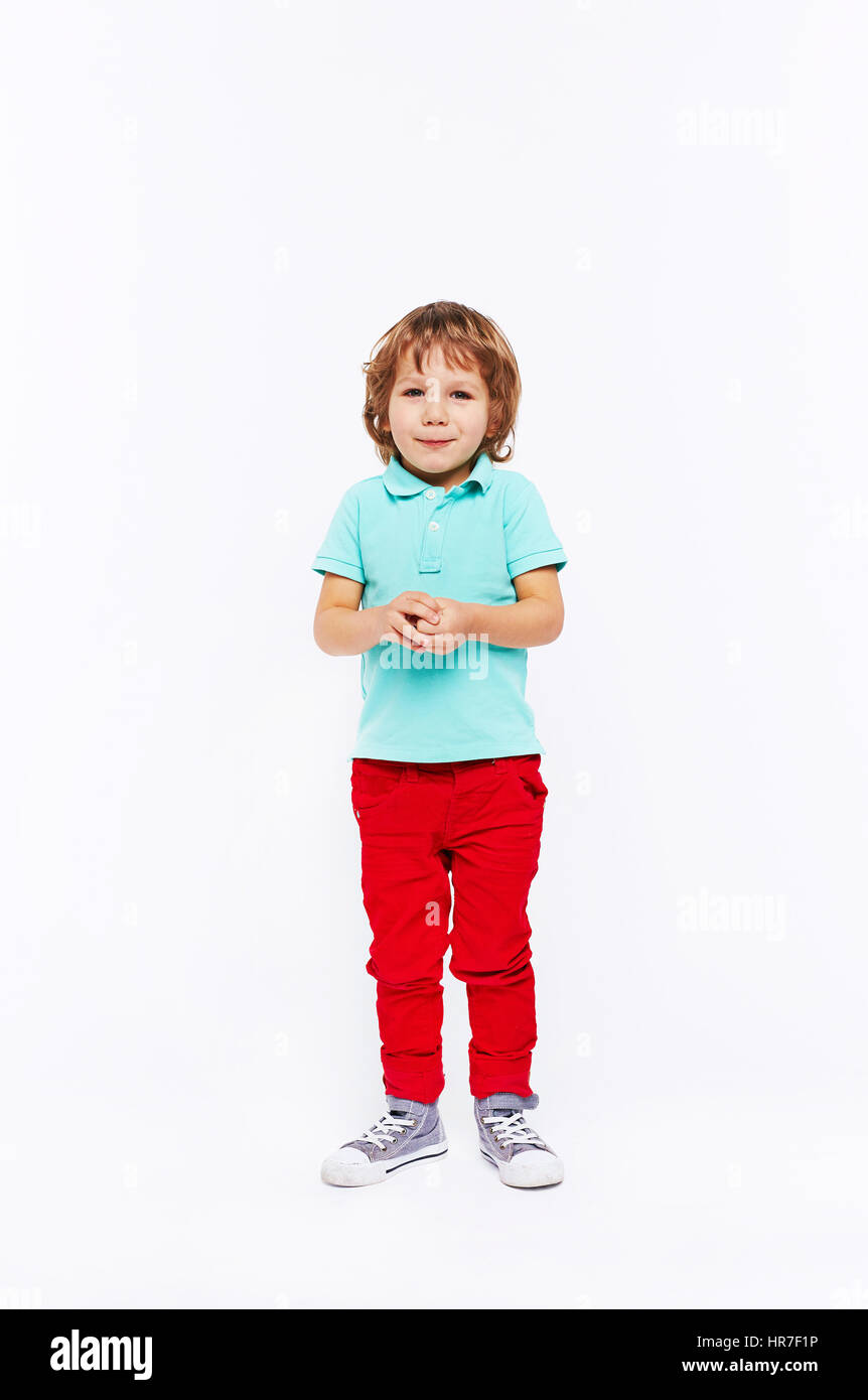 Corpo pieno colpo di timido bambino vestito in bright abiti colorati in piedi e sorridente twiddling mani contro uno sfondo bianco Foto Stock