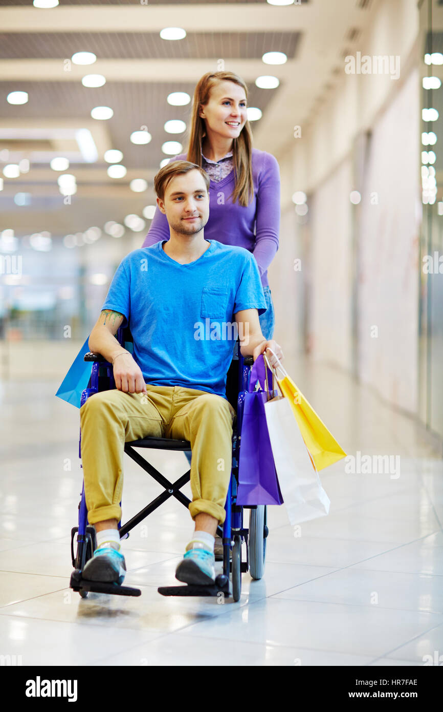 Giovane donna spingendo il suo ragazzo in sedia a rotelle in mall, essi guardando vetrine e colorato di contenimento di sacchetti di carta Foto Stock