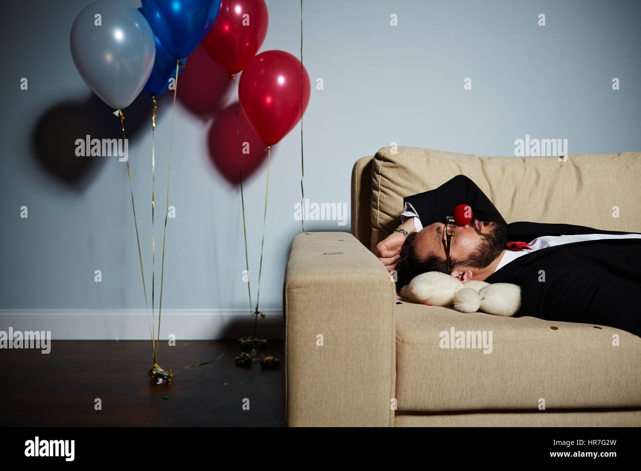 Vita-up ritratto di uomo barbuto con red clown naso che dorme sul divano dopo la festa selvaggia, orso bianco giacente sotto la sua testa Foto Stock