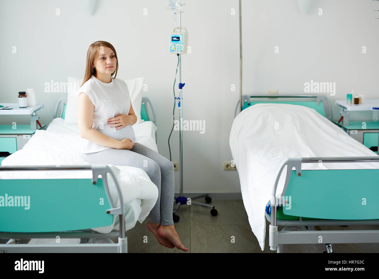 Giovane donna incinta seduta sul letto in confortevoli ward, attesa per medico e accarezzare il suo ventre premurosamente Foto Stock