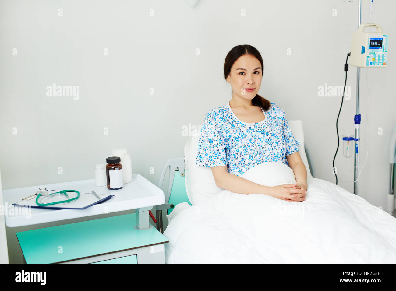 Ritratto di giovane donna incinta giacenti nel letto in confortevoli ward, in attesa di visite ginecologiche check-up e sorridente felicemente Foto Stock