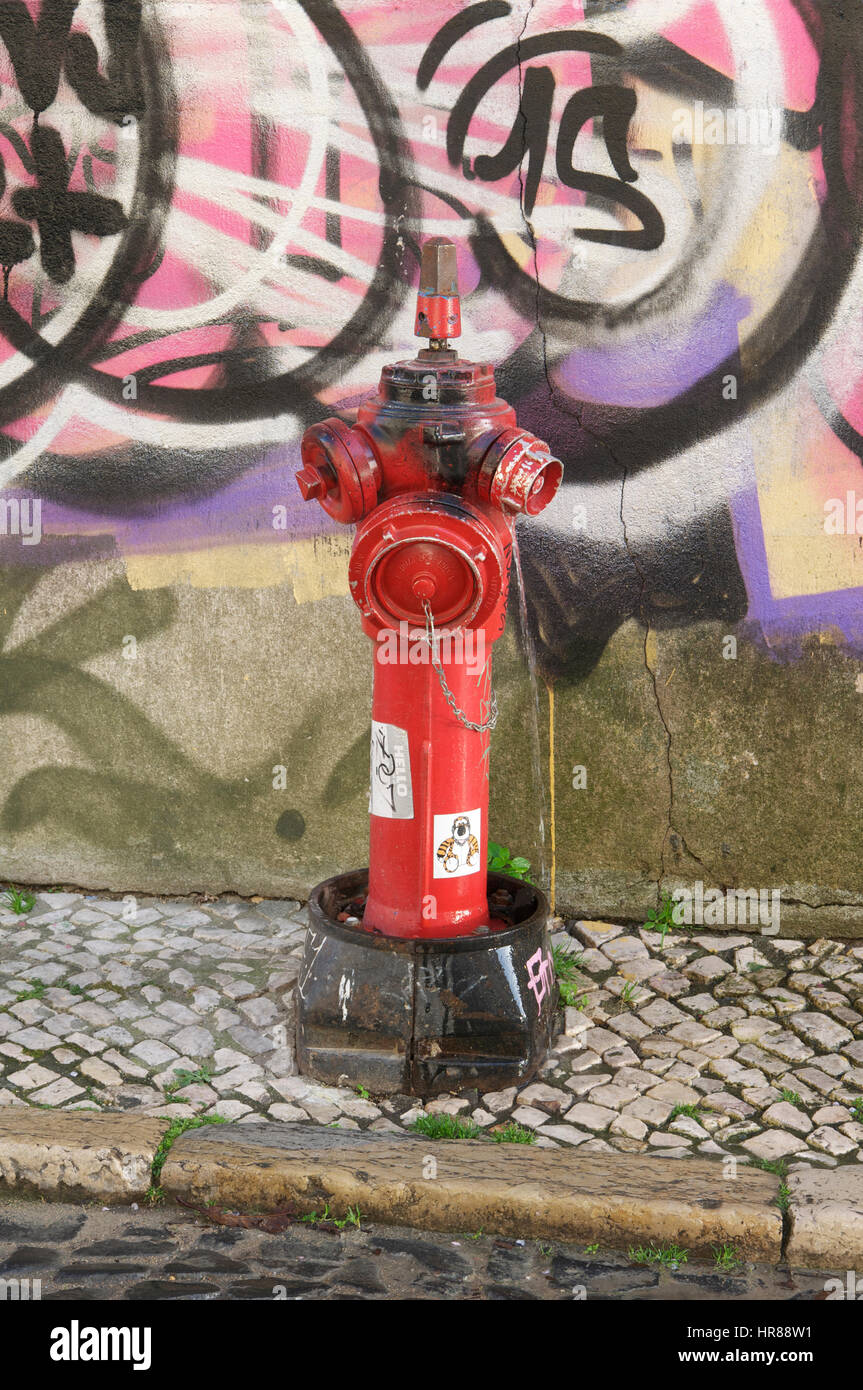 Arredo urbano. Un rosso di emergenza incendio si erge contro un coperto di graffiti nel muro di un ciottolo backstreet di Lisbona, città capitale del Portogallo. Foto Stock